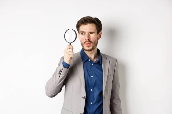 Modelo masculino guapo en traje mirando a través de lupa con interés, viendo algo a un lado, fondo blanco —  Fotos de Stock