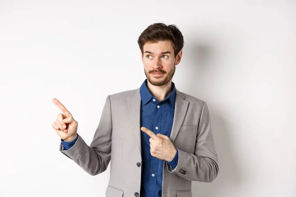 Dubbio imprenditore maschio in giacca e cravatta guardando e indicando a sinistra con il viso esitante, in piedi incerto sullo sfondo bianco — Foto Stock