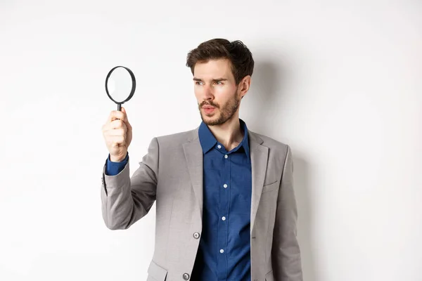 Serious man in suit searching for clues through magnifying glass, investigating, standing on white background — Stock Photo, Image