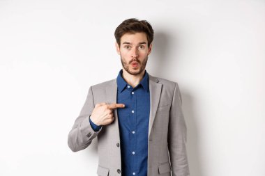 Kim ben mi? Takım elbiseli şaşırmış bir adam kendini inançsızlıkla işaret ediyor, kameraya bakıyor, beyaz arka planda duruyor.