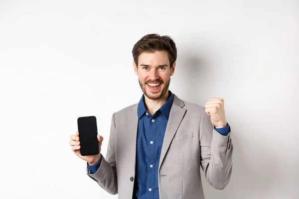 E-commerce et concept d'achat en ligne. Homme faisant de l'argent sur Internet, montrant l'écran du smartphone et le geste du gagnant, souriant satisfait, debout sur fond blanc — Photo