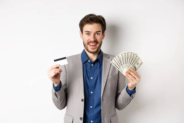 Empresário bem sucedido feliz em terno mostrando dinheiro e cartão de crédito, sorrindo para a câmera, fundo branco — Fotografia de Stock