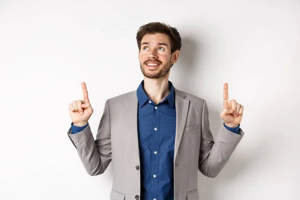 Dreamy smiling male manager in suit pointing and looking up, happy face, checking out advertisement, standing against white background — Stock Photo, Image