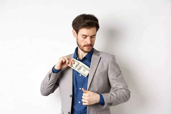 Empresário bem sucedido colocar dólares dentro do bolso terno, escondendo dinheiro, de pé sobre fundo branco — Fotografia de Stock