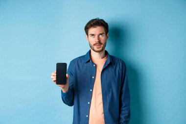 Sinirli genç adam boş akıllı telefon ekranı gösteriyor ve gözleri yuvarlıyor. Mavi arka planda duruyor.