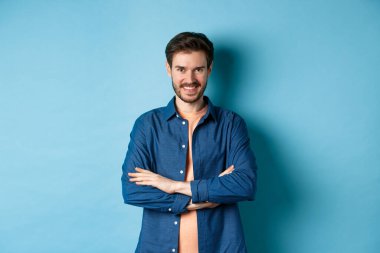 Kendine güveni tam genç bir adam kendini profesyonel hissediyor, kollarını göğsünde kavuşturuyor ve sırıtıyor, mavi arka planda duruyor..