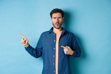Şok olmuş ve gücenmiş genç adam kaşlarını çatmış, yaralı görünüyor, boş alanı işaret ediyor, bir şeyden şikayet ediyor, mavi arka planda duruyor.