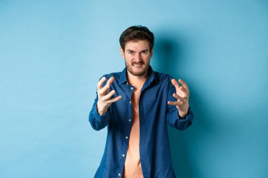 Öfkeli adam birini boğmak ister, düşmanını boğmak için ellerini uzatır, dudaklarını ısırır ve öfkeli görünür, mavi arka planda durur.