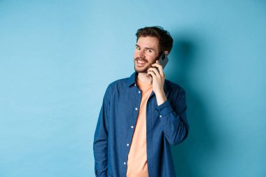 Modern gülümseyen adam cep telefonuyla konuşuyor, kameraya mutlu görünüyor, mavi arka planda duruyor.
