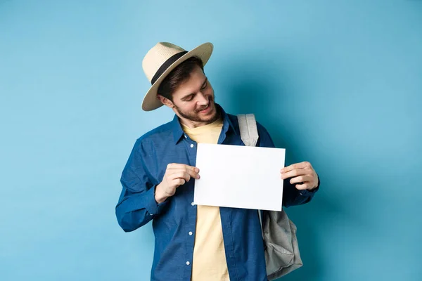 Felice turista in cappello di paglia sorridente e guardando un pezzo di carta per il vostro logo, in piedi su sfondo blu — Foto Stock