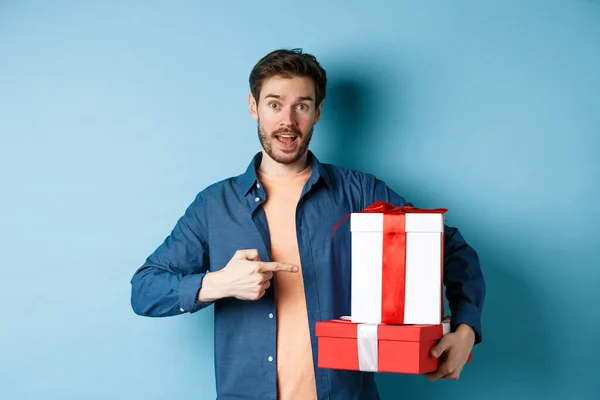 Красивый молодой человек указывает на валентинки подарочные коробки, стоя на синем фоне в повседневной одежде — стоковое фото