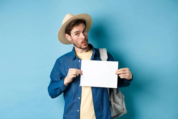 Turista excitado en sombrero de paja, autostop, mostrando un pedazo de papel en blanco y mirando divertido, de pie sobre fondo azul — Foto de Stock