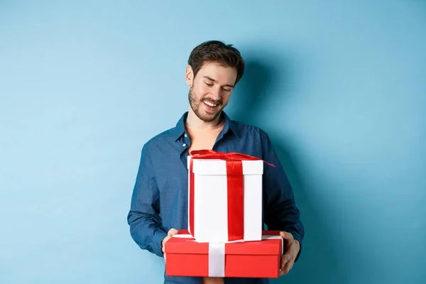Романтичный молодой человек с бородой, счастливо смотрящий на подарочные коробки на День Святого Валентина, дарящий подарки любовнику, стоящий на синем фоне — стоковое фото
