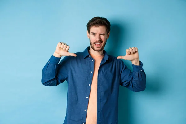 Zelfverzekerde man die knipoogt en naar zichzelf wijst, zichzelf promoot, op een blauwe achtergrond staat — Stockfoto