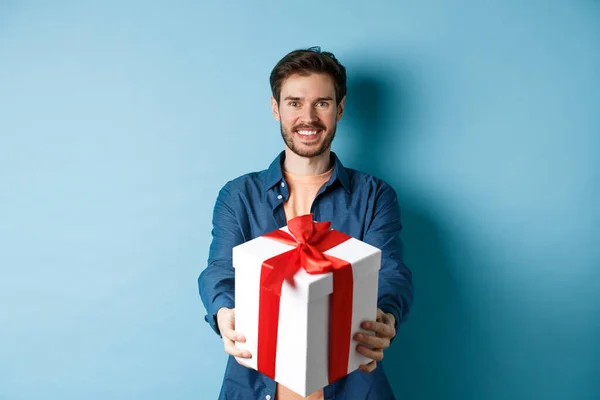День Святого Валентина. Красивый улыбающийся мужчина протягивает руку с подарочной коробкой, желая счастливого праздника. Парень делает сюрприз подарок и выглядит веселым, стоя на синем фоне — стоковое фото
