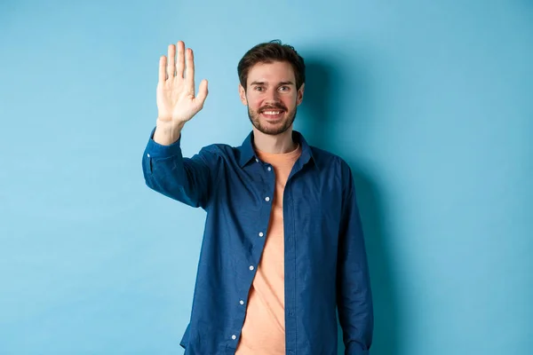 Amistoso joven sonriendo y levantando la mano para los cinco altos, renunciando a saludar o decir hola, de pie sobre fondo azul — Foto de Stock