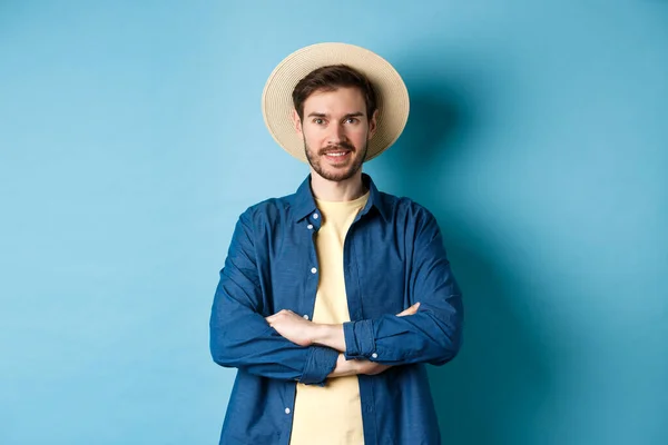 穿着草帽外出度假、胸前交叉双臂、面带微笑、看起来自信、站在蓝色背景上的快乐男人的画像 — 图库照片
