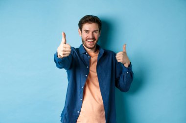 Olumlu gülümseyen adam başparmağını kaldırıyor ve gülümsüyor, sana iltifat ediyor, iyi iş çıkartıyor, iyi hareket ediyor, mavi arka planda duruyor.