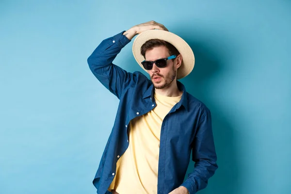 Chico fresco que se divierte en vacaciones, usando sombrero de paja y gafas de sol, mirando a un lado atrevido, de pie sobre fondo azul — Foto de Stock