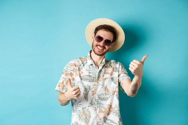 Turista alegre se divertindo férias de verão, mostrando polegares para cima e sorrindo, de pé em camisa havaiana e óculos de sol no fundo azul — Fotografia de Stock