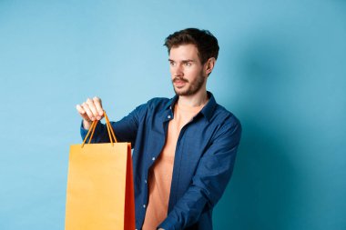 Yakışıklı genç adam, alışveriş satıcısı müşteriye turuncu alışveriş torbası uzatıyor, yan bakıyor, mavi arka planda duruyor.