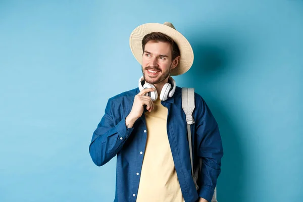 Turista guapo con mochila y sombrero de paja mirando a un lado, sonriendo y tocando auriculares, de pie sobre fondo azul — Foto de Stock