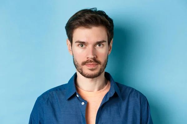 Närbild av ung man med skägg tittar allvarligt på kameran, står i ledig skjorta på blå bakgrund — Stockfoto
