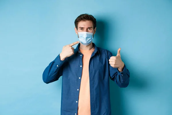 Coronavírus e conceito de pandemia. Jovem homem saudável apontando para máscara médica e mostrando os polegares para cima, usando medidas preventivas de pegar covid-19, fundo azul — Fotografia de Stock