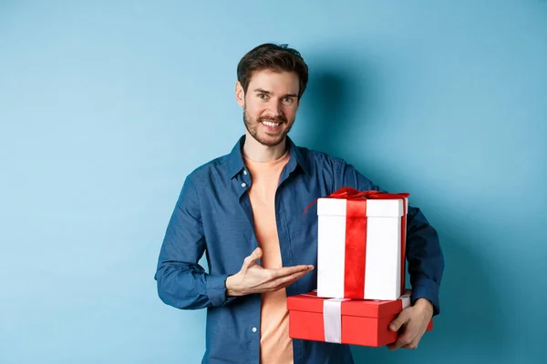 Молодой человек в повседневной одежде покупает романтические подарки на День Святого Валентина, указывая на коробки подарков и улыбаясь, стоя на синем фоне — стоковое фото