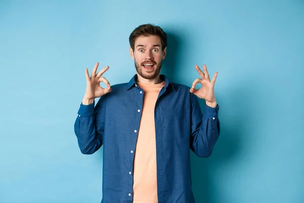 Hombre impresionado alabando algo impresionante, mostrando signos de bien y mirando sorprendido, elogiando lo bueno, de pie sobre fondo azul — Foto de Stock