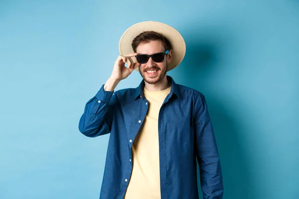 Feliz chico sonriente que va de vacaciones de verano, con sombrero de paja y gafas de sol negras, de pie sobre fondo azul — Foto de Stock