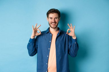 Tatmin olmuş genç adam gülümsüyor ve iyi bir jest yapıyor, iyi bir şeyi onaylıyor, mükemmel bir şeyi övüyor, mavi arka planda duruyor.