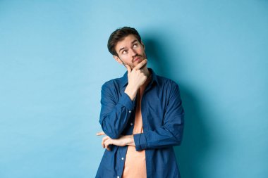 Düşünceli genç adam yüzüne dokunuyor ve boş alana bakıyor, bir şeyler hayal ediyor, plan dışı düşünüyor, mavi arka planda duruyor.