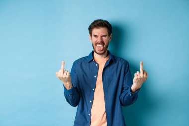 Ignorant and rude guy showing middle finger and smiling, say fuck you, standing on blue background