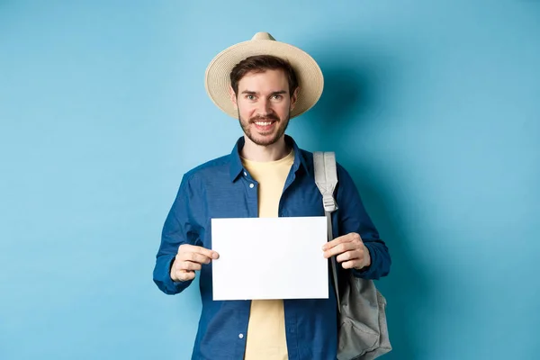 Turista feliz con mochila mostrando un pedazo de papel vacío, sonriendo a la cámara, de pie sobre fondo azul. Concepto de vacaciones de verano y vacaciones — Foto de Stock