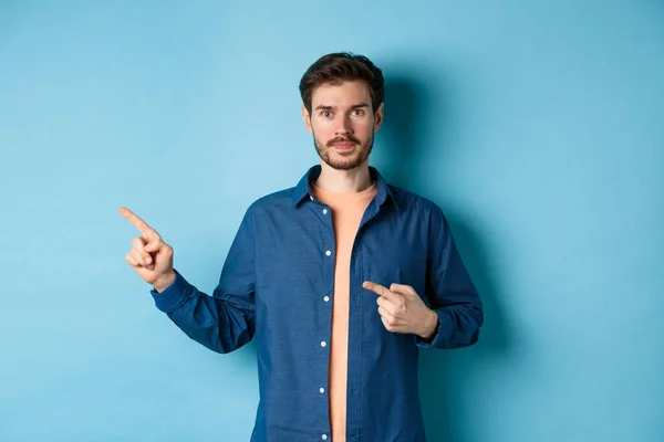 Bello giovanotto in camicia, punta il dito verso sinistra al promo, mostrando il logo, in piedi su sfondo blu — Foto Stock