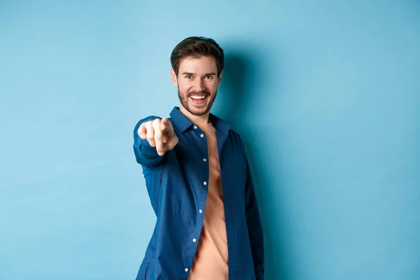 Zelfverzekerde lachende man die jou kiest of uitnodigt, wijzend naar camera beslissend, staand op blauwe achtergrond — Stockfoto