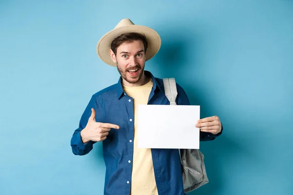 Turista guapo sonriendo y señalando un pedazo de papel vacío, usando sombrero de paja y mochila. Concepto de vacaciones de verano — Foto de Stock