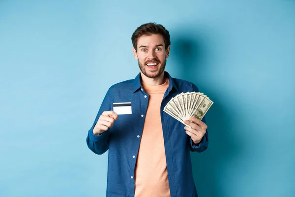 Sonriente chico caucásico mostrando tarjeta de crédito de plástico y dinero, mirando feliz, de pie sobre fondo azul — Foto de Stock