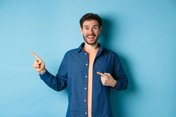 Eccitato e felice uomo caucasico sorridente alla macchina fotografica, indicando il dito sinistro allo spazio vuoto, mostrando il logo, in piedi su sfondo blu — Foto Stock