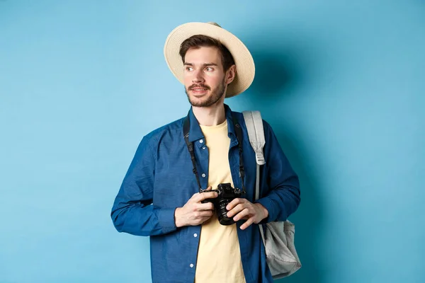 Guapo chico positivo en sombrero de verano, la celebración de la cámara de fotos y mirando a un lado, turista tomando fotos de vacaciones, de pie con mochila sobre fondo azul — Foto de Stock