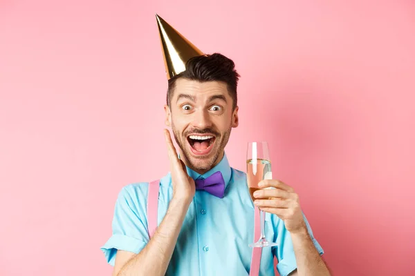 Vakantie en feest concept. Grappige jongeman in verjaardagshoed viert feest, schreeuwt van vreugde en verrassing, heft glas champagne en glimlacht, staat op roze achtergrond — Stockfoto