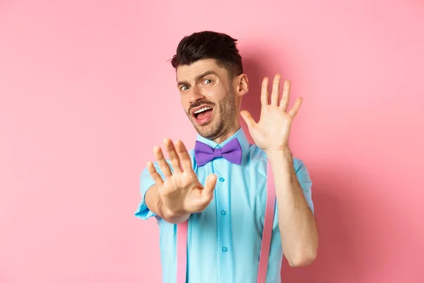 Joven tonto con pajarita y tirantes pidiendo parar, cubriendo la cara con las manos y gritando aterrorizado, mirando algo asqueroso, de pie sobre fondo rosa —  Fotos de Stock