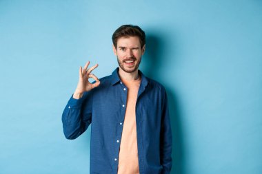 Olumlu gülümseyen adam göz kırpıyor ve iyiye işaret gösteriyor, iyi işi övüyor, iyi bir arkadaş tavsiye ediyor, mavi arka planda duruyor.