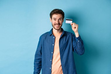 Mutlu beyaz adam gülümsüyor, plastik kredi kartı gösteriyor, mavi arka planda sıradan kıyafetler giyiyor..
