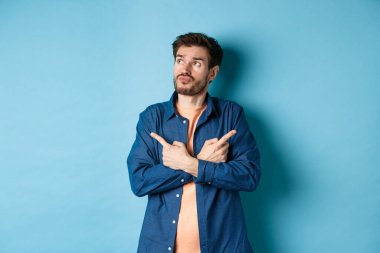 Kararsız sakallı adam çapraz kollar ve iki farklı türü gösteren, seçim yapan, mavi arka planda duran.