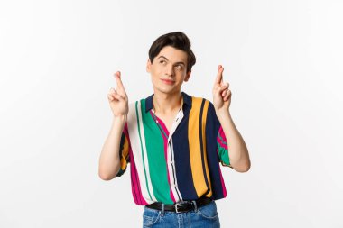 Hayalperest genç adam parmaklarını çaprazlayarak dilek diliyor, gülümsüyor ve sol üst köşeye bakıyor düşünceli, beyaz arka planda duruyor..