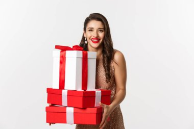 Yeni yıl, Noel ve kutlama konsepti. Parti elbisesi içinde güzel bir kadın, hediyeler ve hediyeler taşıyor, mutlu bir şekilde gülümsüyor, beyaz arka planda duruyor.