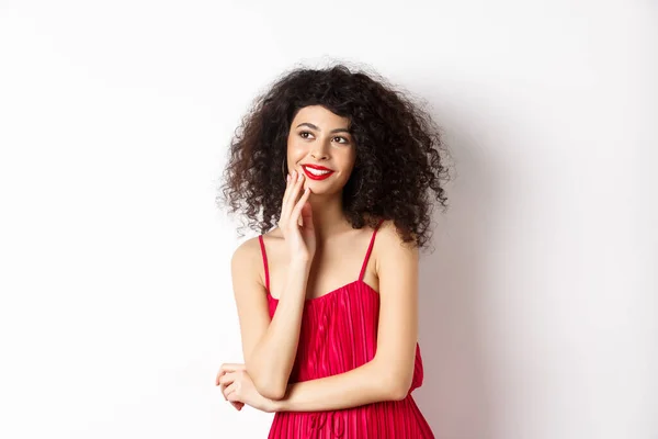 Menina elegante sonhador com cabelo encaracolado, vestindo vestido vermelho e maquiagem, olhando para a esquerda e sorrindo para o logotipo, de pé sobre fundo branco — Fotografia de Stock