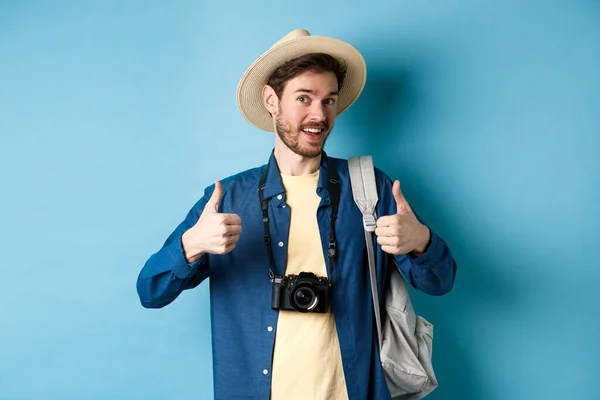 Buon turista sorridente dire di sì e mostrare pollici in su, andando in vacanza estiva, tenendo zaino e macchina fotografica, sfondo blu — Foto Stock